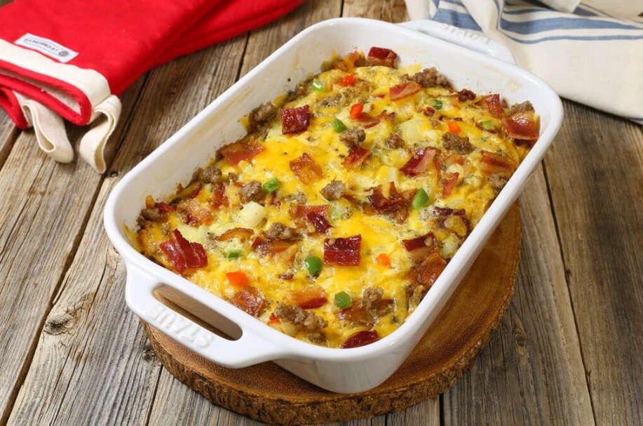 A baked breakfast casserole in a white dish on a wooden surface, featuring melted cheese, bacon, and vegetables. Nearby, a red oven mitt adds a vibrant touch.
