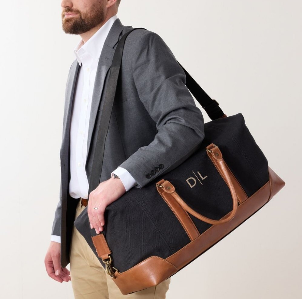 A man in a suit carries a black and tan duffel bag, standing confidently in a professional setting.