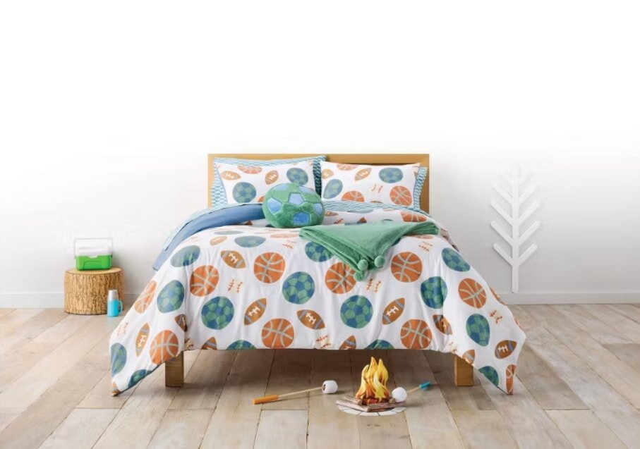 Cozy bed with sports-themed bedding featuring basketball and soccer motifs, topped with a green throw. Nearby, marshmallows on sticks by a small campfire create a playful, camping vibe.