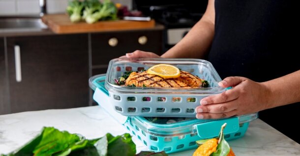 A person holds a turquoise container with grilled salmon topped with lemon, placed on a kitchen counter. Fresh vegetables and another container are visible nearby, creating a fresh and organized cooking scene.