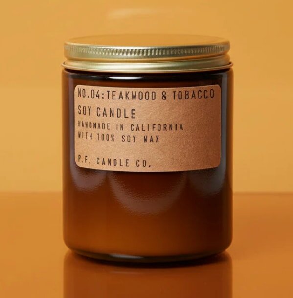 Amber jar candle labeled "Teakwood & Tobacco," handmade in California with soy wax, by P.F. Candle Co., on orange background.
