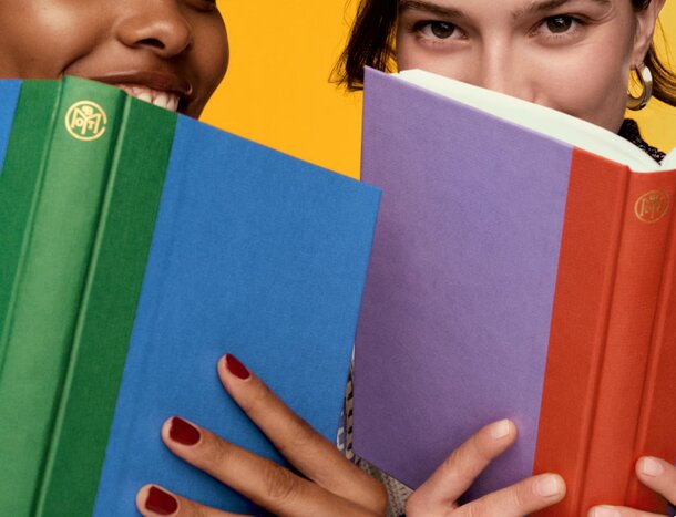 Two women stand against a yellow background, each holding colorful books and smiling at the camera.