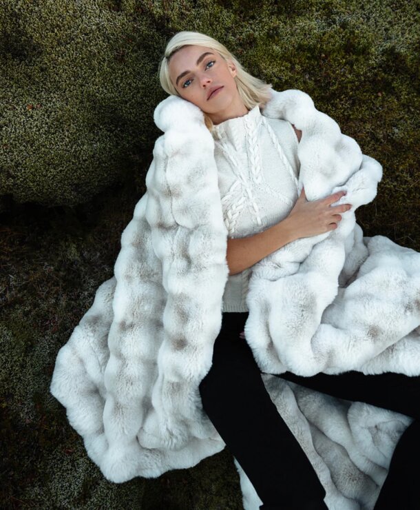 A woman reclines on a rock, wrapped in a cozy fur blanket, enjoying a serene outdoor moment.
