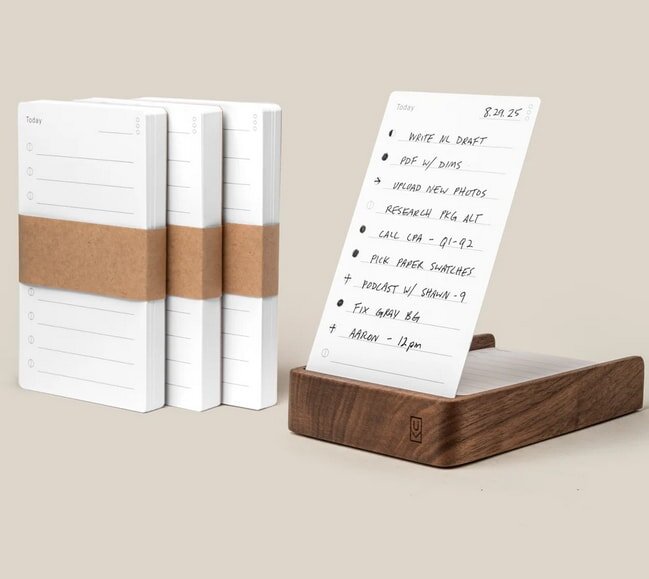 Wooden stand holding a to-do list card with tasks. Three wrapped notepad stacks are beside it on a beige background.