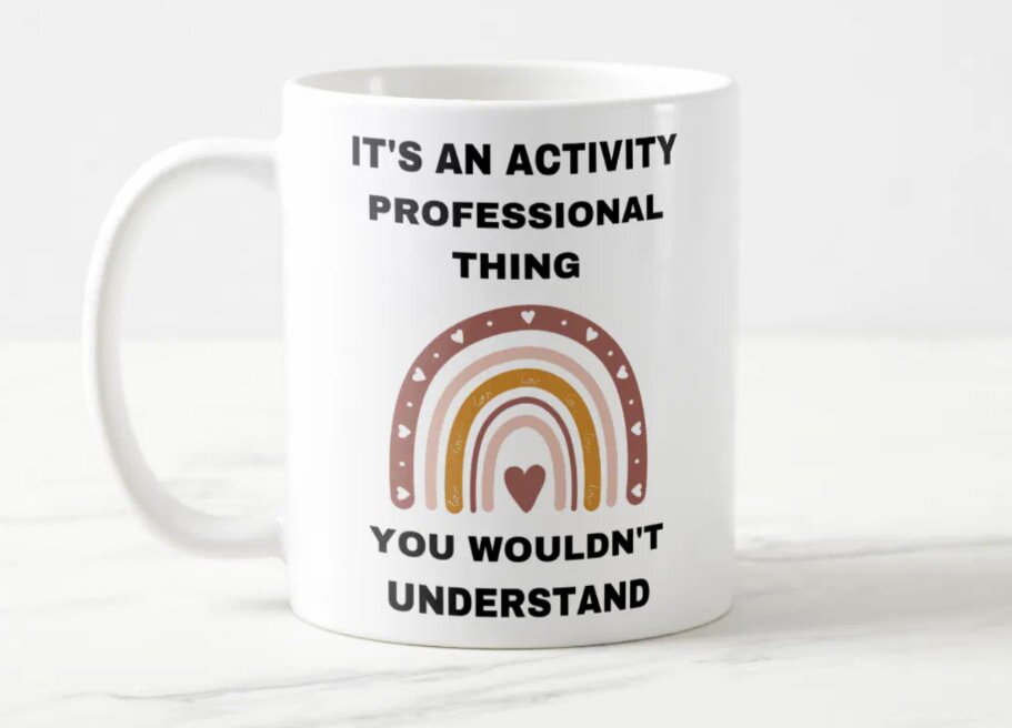 White mug featuring a multi-colored rainbow with hearts. Above, bold text says, "It's an activity professional thing." Below, "You wouldn't understand."