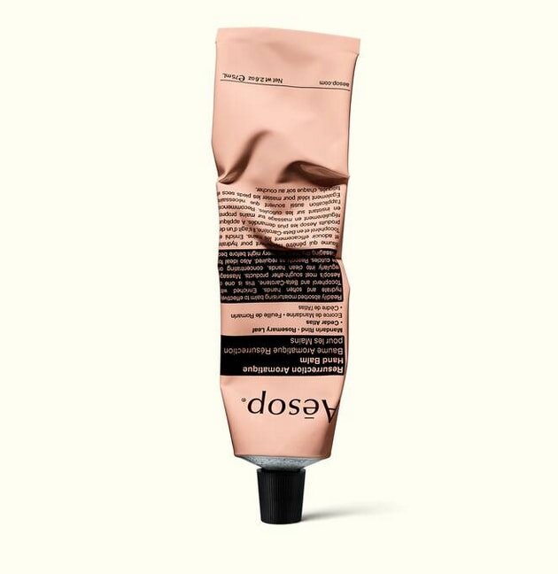 A crumpled pink tube of Aesop hand balm lies against a plain background, showcasing its minimalist design and black cap.