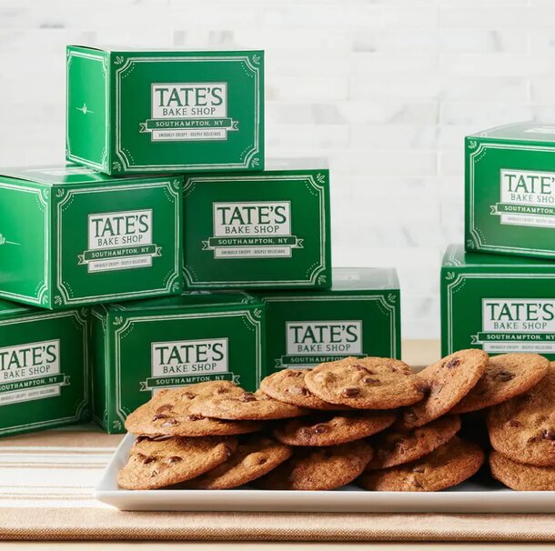 Stacks of green Tate’s Bake Shop boxes are in the background with a plate of golden-brown chocolate chip cookies in the foreground, conveying a sense of warmth and indulgence.
