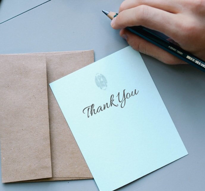 A person's hand holds a pencil near a light blue card with "Thank You" written on it. A brown envelope lies underneath, conveying gratitude and warmth.