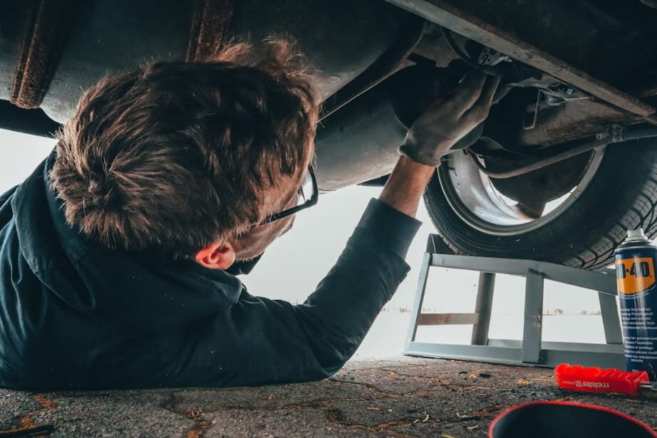 A man is working underneath a car, focused on repairs while positioned near the vehicle's wheels.