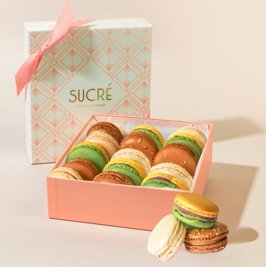 A pastel pink box of assorted macarons in various colors like green and brown sits open beside a patterned gift box with a pink ribbon. Luxurious and inviting.