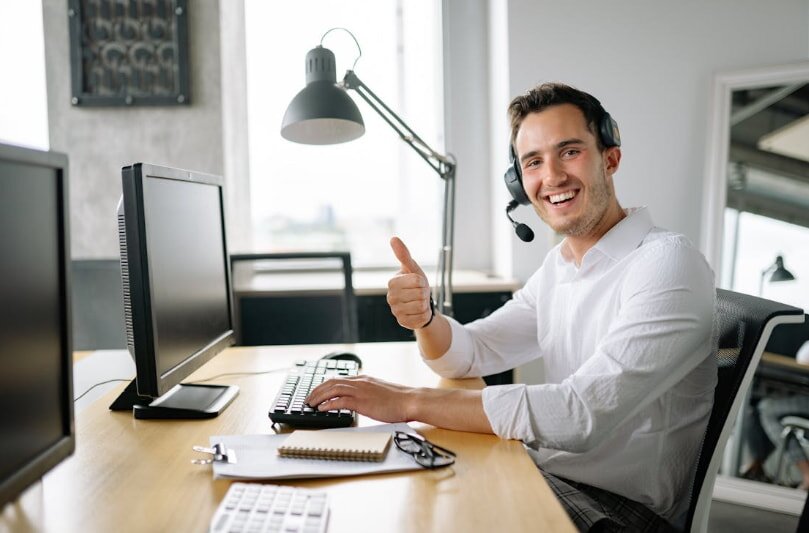 A man in a white shirt sits at a desk, working on a computer with a phone beside him.
