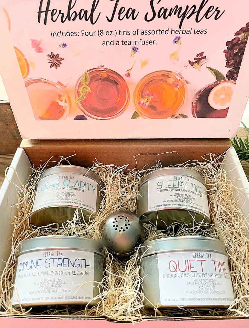 A pink box labeled "Herbal Tea Sampler" contains four tins of herbal tea labeled "Clear Clarity," "Sleepy Time," "Immune Strength," and "Quiet Time," nestled in straw with a metal tea infuser. The box conveys a calming and inviting tone.