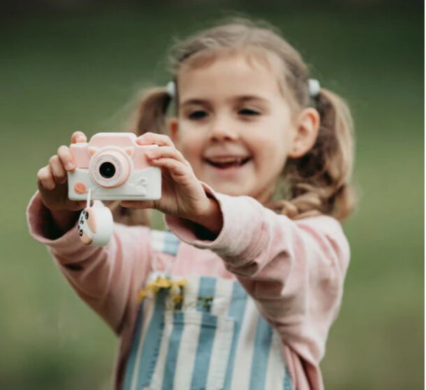 A young girl joyfully holds a pink camera, ready to capture moments with a bright smile on her face.