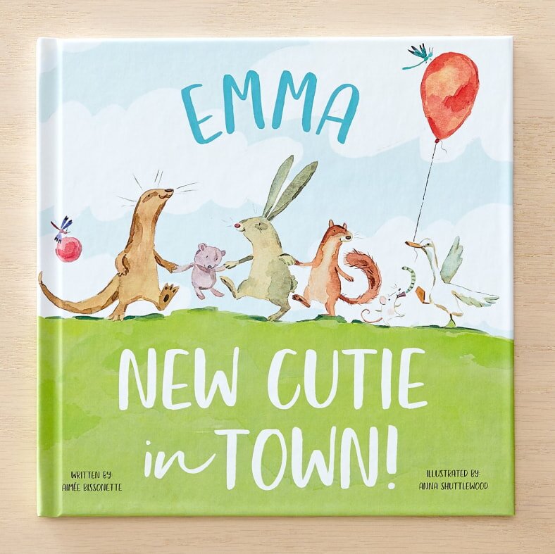 Children's book cover titled "Emma: New Cutie in Town." Features joyful animals and a balloon on a grassy hill under a bright sky. Cheerful and welcoming.