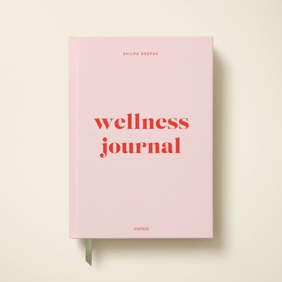 A pink wellness journal with "wellness journal" in bold red text on the cover. A subtle green ribbon bookmark peeks out from the bottom.