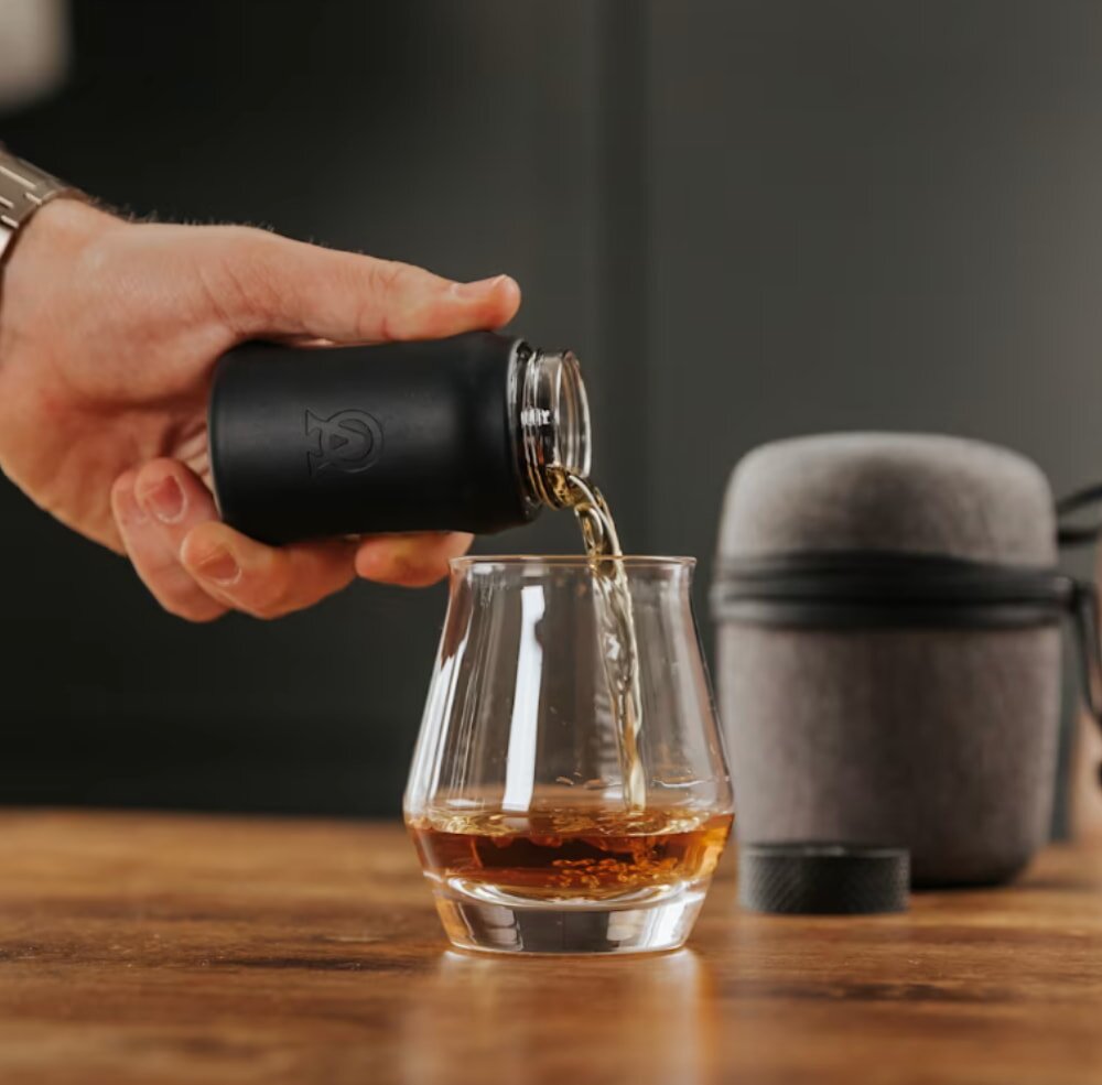 A person pouring whisky from a bottle into a clear glass on a wooden table.