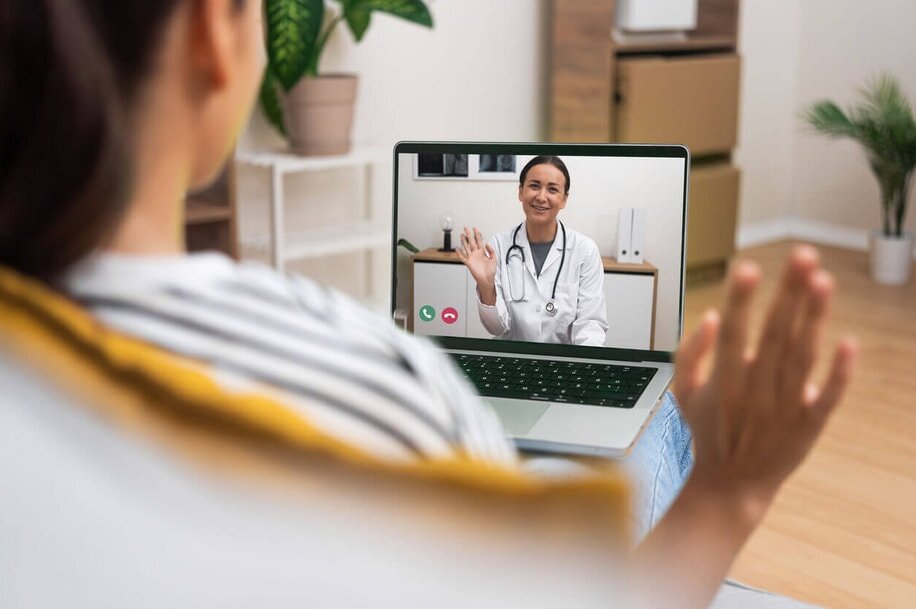 A woman sits on a couch with her laptop, video chatting with a doctor displayed on the screen.