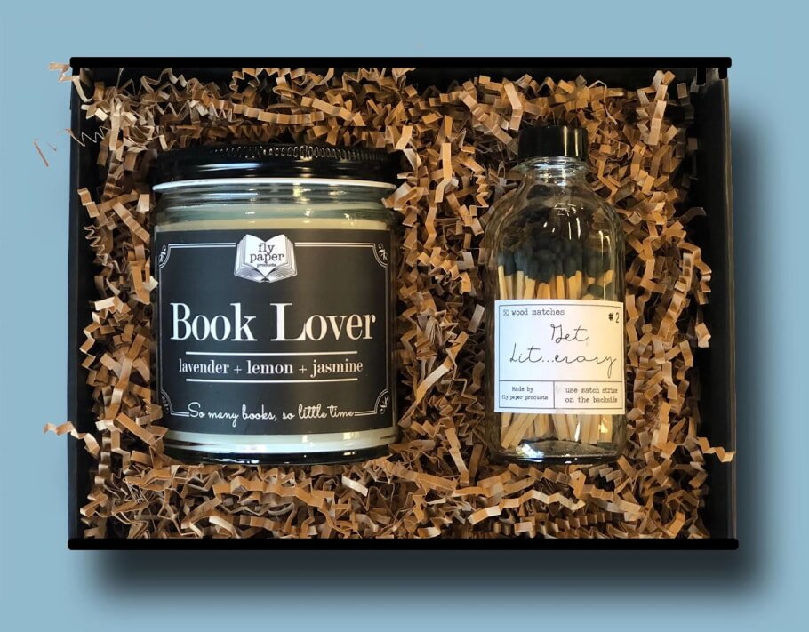 A gift box contains a "Book Lover" candle and a jar of matches, nestled in brown crinkle paper. The candle label shows lavender, lemon, and jasmine scents.