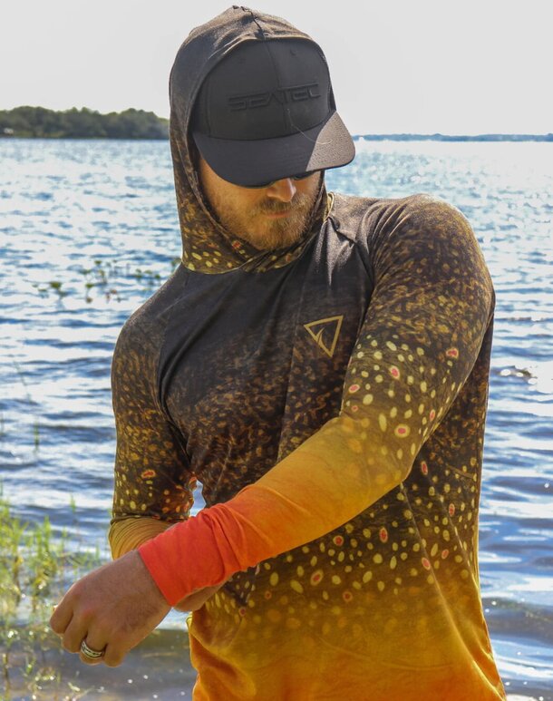 A man wearing a hat and a fish-patterned shirt, smiling outdoors.