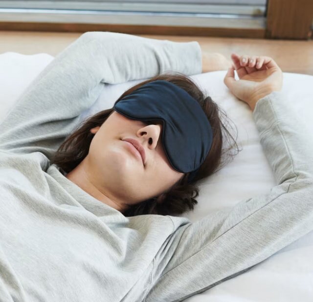 A woman wearing a gray shirt sleeps peacefully with a black eye mask, lying on white sheets, arms relaxed above her head, conveying tranquility.
