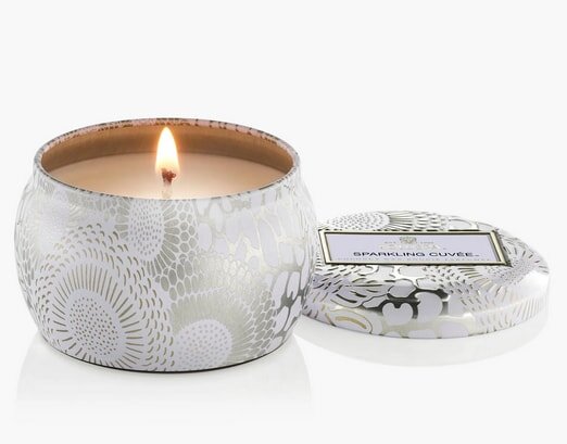 Decorative tin candle labeled "Sparkling Cuvée" with a lit flame, featuring a floral pattern and an open lid beside it.
