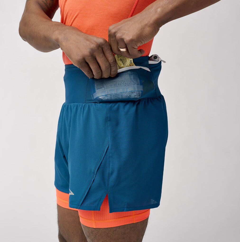A man in an orange shirt and blue shorts holds his wallet, standing casually outdoors.