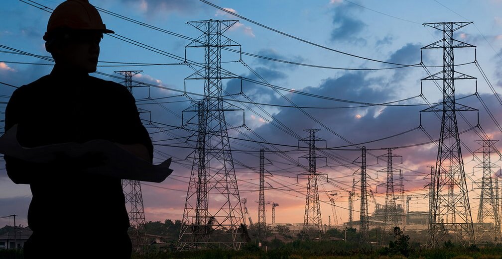 Silhouette of a person in a hard hat holding plans, standing before power lines and towers at dusk. The scene conveys a focus on energy and construction.