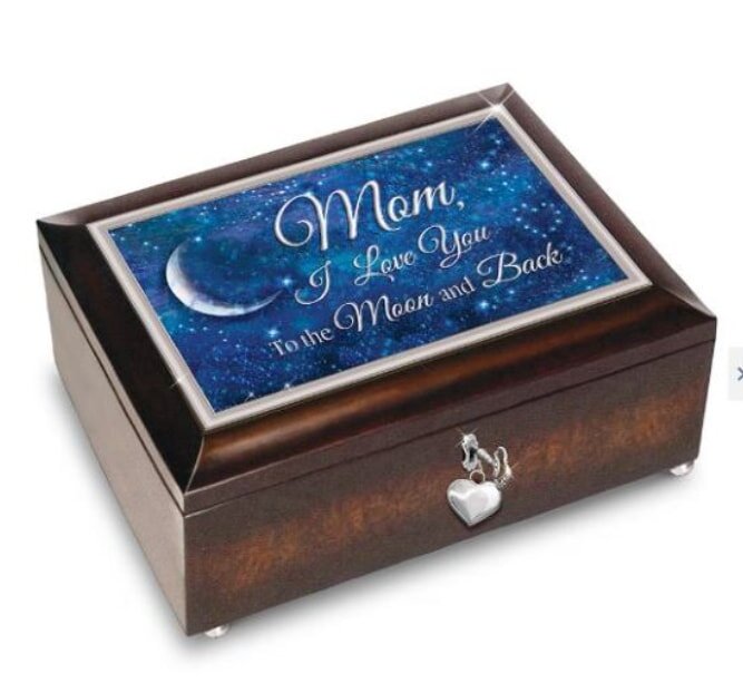 A dark wooden jewelry box with a silver heart-shaped latch. The lid has a starry sky design with a crescent moon and the words, "Mom, I Love You to the Moon and Back" in elegant script. The tone is sentimental and loving.