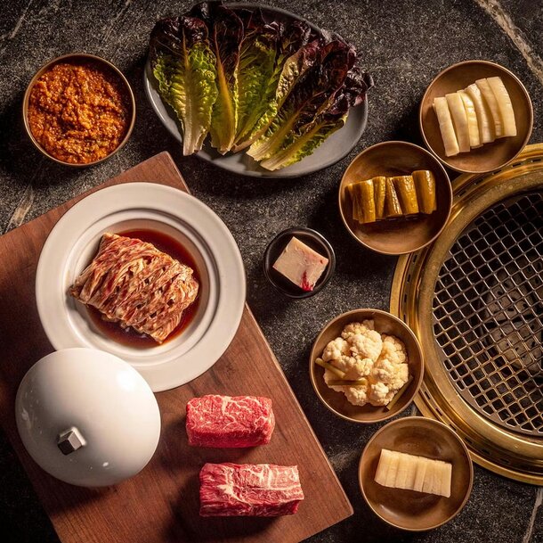A selection of Korean barbecue ingredients is artfully arranged on a rustic table. Fresh lettuce, marinated beef, kimchi, and various pickled vegetables surround a grill, evoking a warm, inviting dining experience.