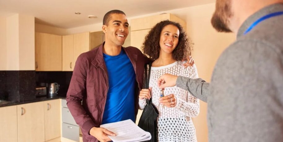 A smiling couple stands in a kitchen holding documents, engaging with a person in a gray shirt. The setting is bright and suggests a positive interaction.