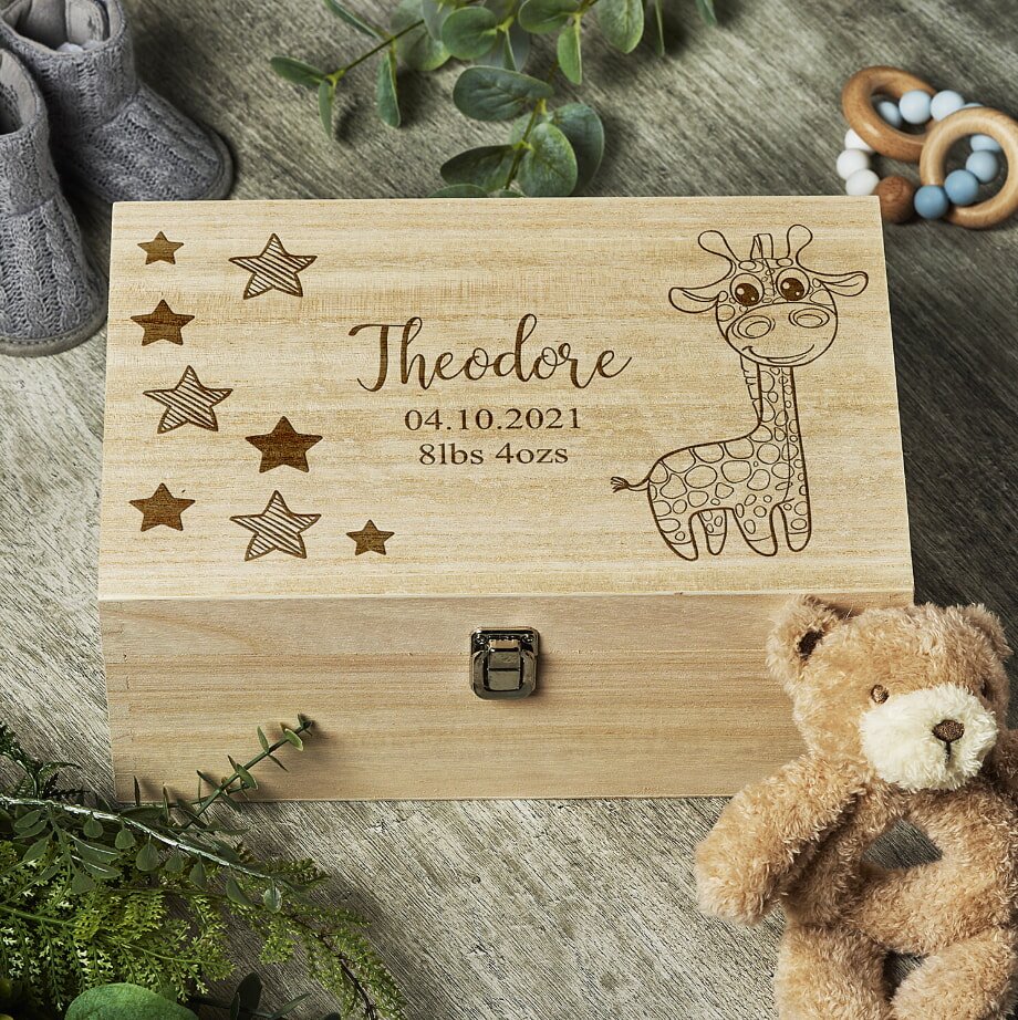 A wooden keepsake box engraved with stars, a cartoon giraffe, and "Theodore, 04.10.2021, 8lbs 4ozs." Nearby are baby shoes, a plush teddy bear, and greenery, creating a cozy, welcoming tone.