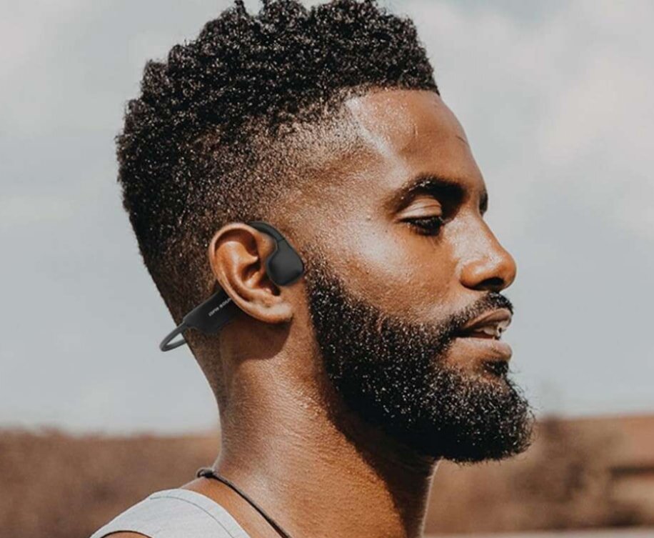 Man outdoors wearing bone conduction headphones, eyes closed, appearing relaxed. Sunny background with a clear sky, conveying a serene mood.