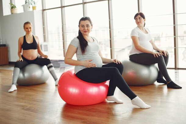Three women are seated on exercise balls in a gym, engaging in a fitness activity together.