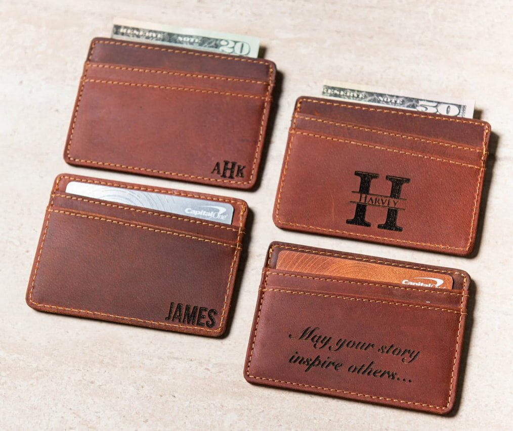 Four brown wallets or cardholders are displayed, each holding a few bills. The wallets have various monograms or names embossed on the front, along with one that features an inspirational phrase.