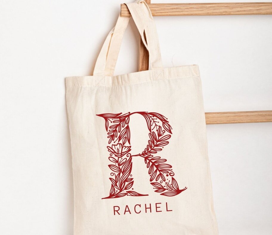 A beige tote bag hangs on a wooden ladder. It features a large, ornate red letter "R" with floral designs, and the name "Rachel" below it in bold.