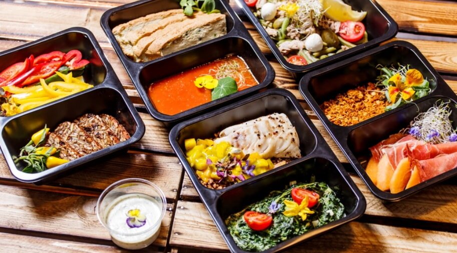 A colorful assortment of gourmet meals in black trays on a wooden surface. Includes salads, grilled meats, vegetables, and bread, evoking a fresh, vibrant feel.