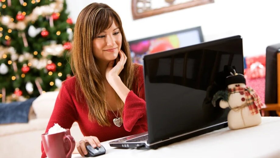 A woman in a red sweater smiles while using a laptop in a cozy setting. A decorated Christmas tree and a snowman plush are in the background, creating a festive atmosphere.