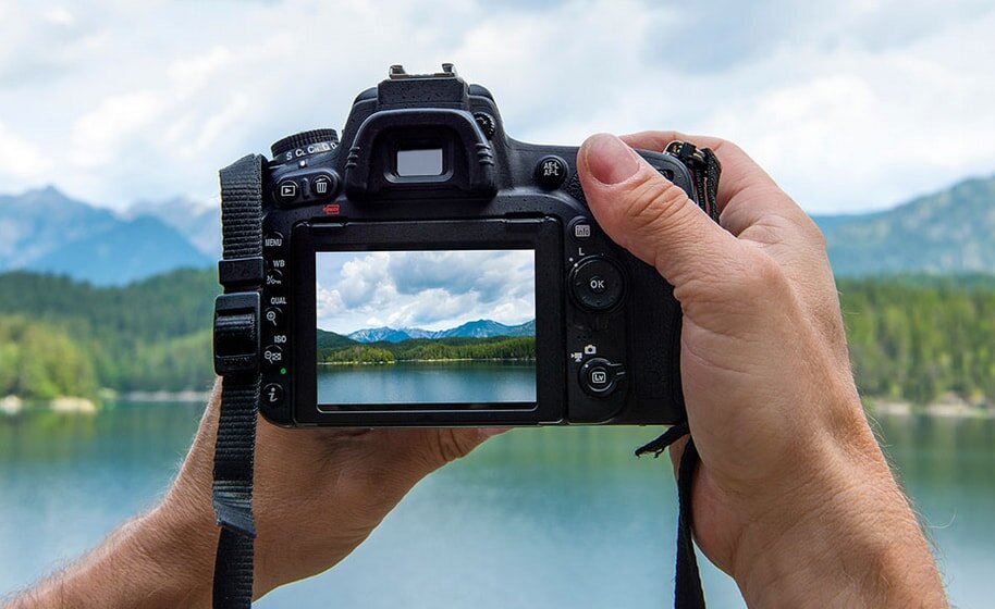 Hands holding a DSLR camera focus its screen on a serene lake and mountains view, capturing nature's beauty. Bright, calm atmosphere with cloudy sky.