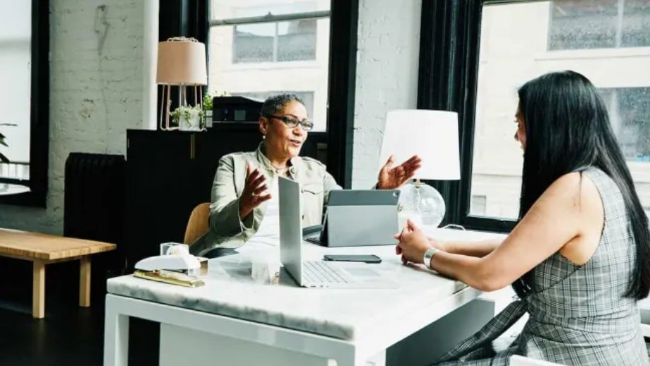 Two women engaged in conversation at a table in a modern office setting.