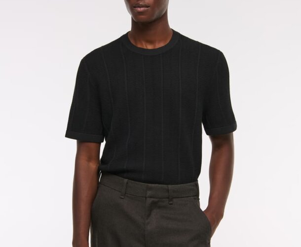 A man wearing a black t-shirt and grey trousers stands confidently, showcasing a casual yet stylish outfit.