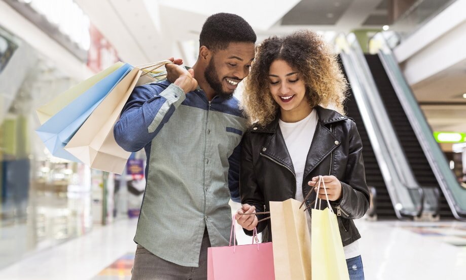 A cheerful couple shopping in a mall; the man holds colorful bags over his shoulder while they both smile and look at more bags. An escalator is visible in the background.