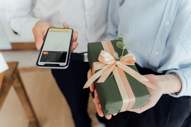A man holds a wrapped gift box in one hand and a smartphone in the other, smiling at the camera.