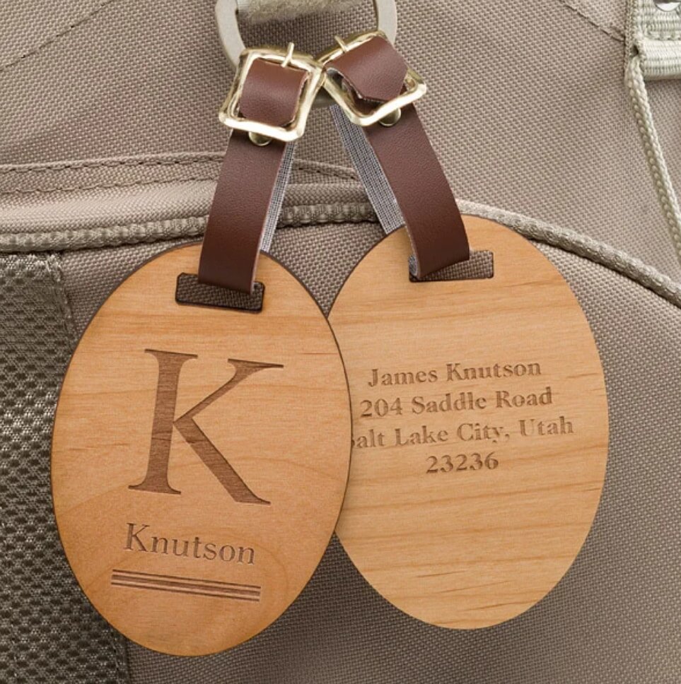 Two oval wooden luggage tags with engraved text hang from brown leather straps on a beige bag. One shows a large "K" and "Knutson," the other has an address.