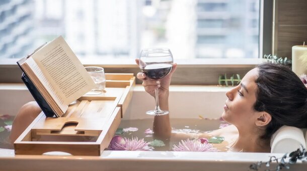A woman relaxes in a bathtub, engrossed in reading a book, surrounded by bubbles and soft lighting.