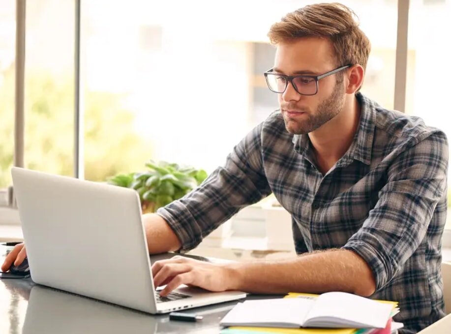 A man in glasses is engaged in work on his laptop, seated at a desk with a notepad nearby.