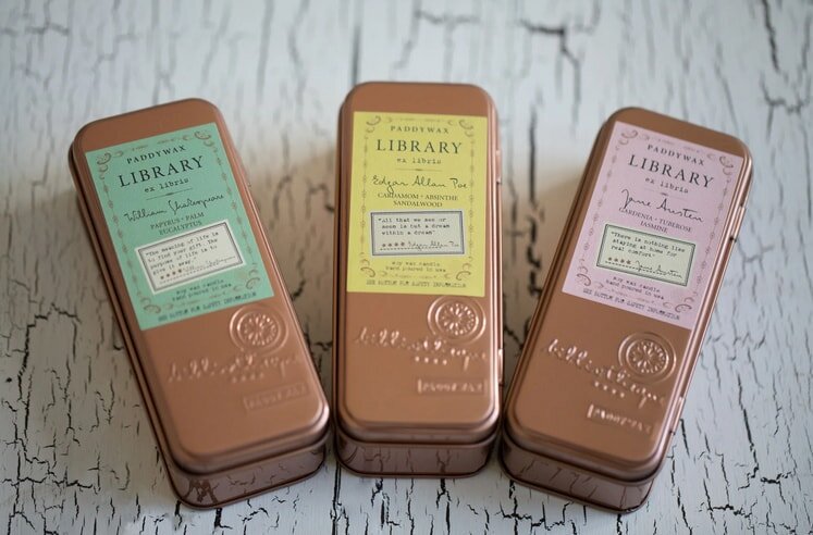 Three Paddywax Library candles in bronze tins featuring Shakespeare, Poe, and Austen on a rustic white wooden surface.