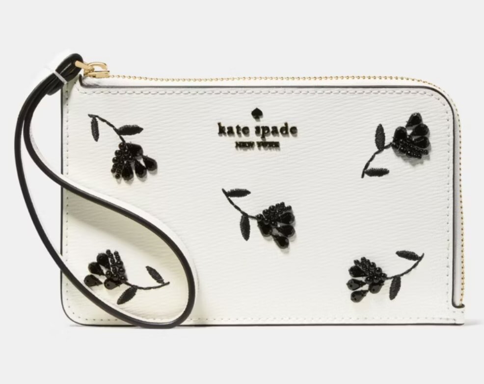 White wristlet with black floral embroidery and "Kate Spade New York" logo. It has a textured finish, gold zipper, and sleek wrist strap.