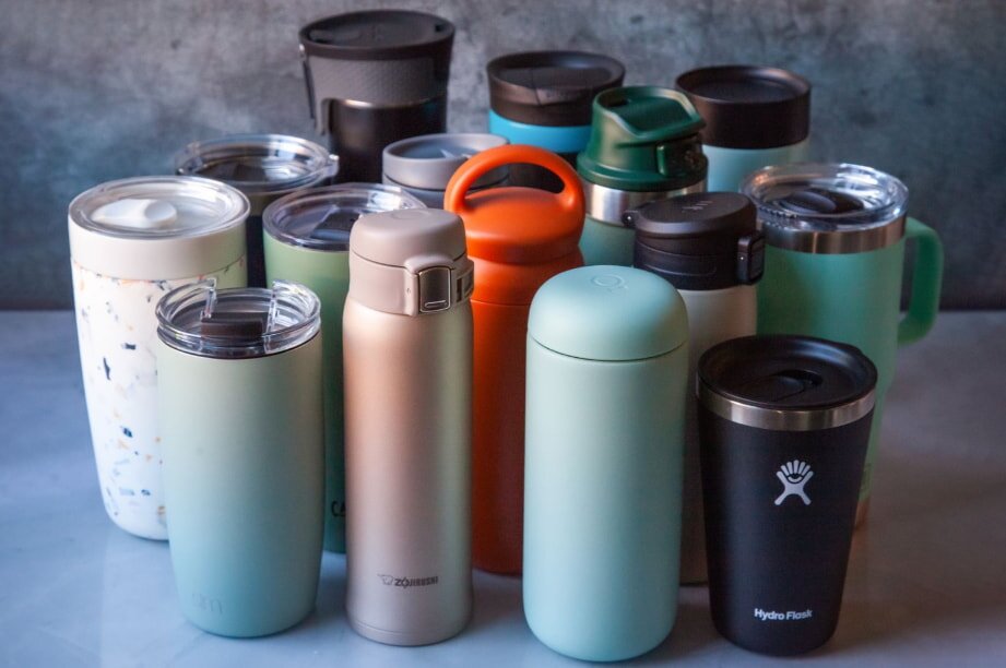 A variety of colorful, reusable travel mugs and bottles are clustered on a table against a gray background. They convey a sense of sustainability and practicality.