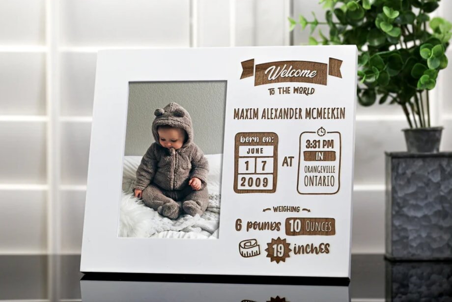 A baby photo frame displays a child in a fuzzy bear outfit on a fluffy white blanket. On the right, birth details in playful brown text. A small potted plant is visible in the background.