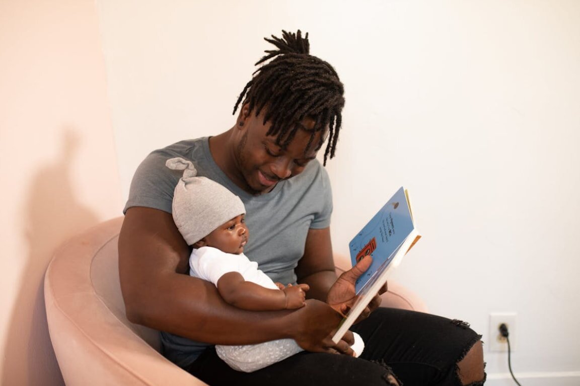 A man sits in a chair, reading a book to his baby, who is nestled comfortably in his lap.